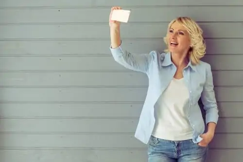 Beautiful mature woman in casual clothes is making a selfie using phone and smiling, standing against gray background